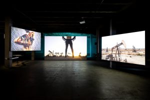 Kolme videoteosta: vasemmalla henkilö hieroo öljyä selkäänsä, keskellä nainen seisoo öljynporausalueella ja oikealla nainen ratsastaa öljypumpun päällä.