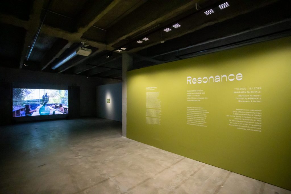 Kuvassa on taidenäyttelytila, jossa on betoninen lattia ja etualalla on vihreä seinä, jossa lukee taidenäyttelyn nimi Resonance ja pienempää tekstiä näyttelystä. Takana näkyy seinälle heijastettu video, jossa nainen laulaa tyhjässä kasvihuoneessa.