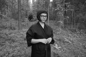 ida fleming portretti taiteilijasta. Mustat vaatteet ja pipo, nainen katsoo alaspäin ja hänen takanaan on metsää. 
