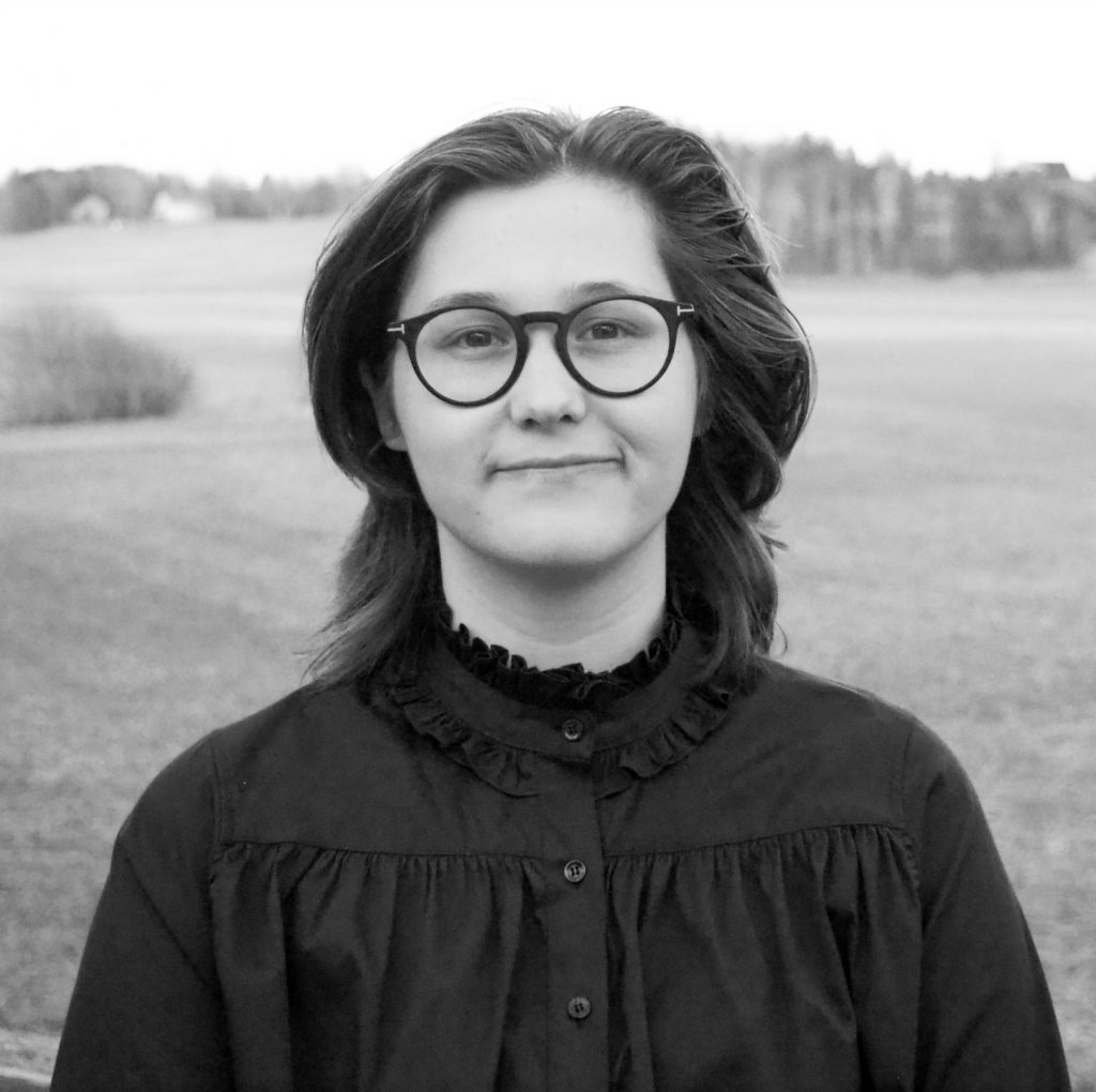 henkilökuva, jossa silmalasinen, puolipitkät hiukset oleva henkilö katsoo suoraan kameraan hymyillen vähän. Kuva on otettu ulkona nurmikkoalueella.