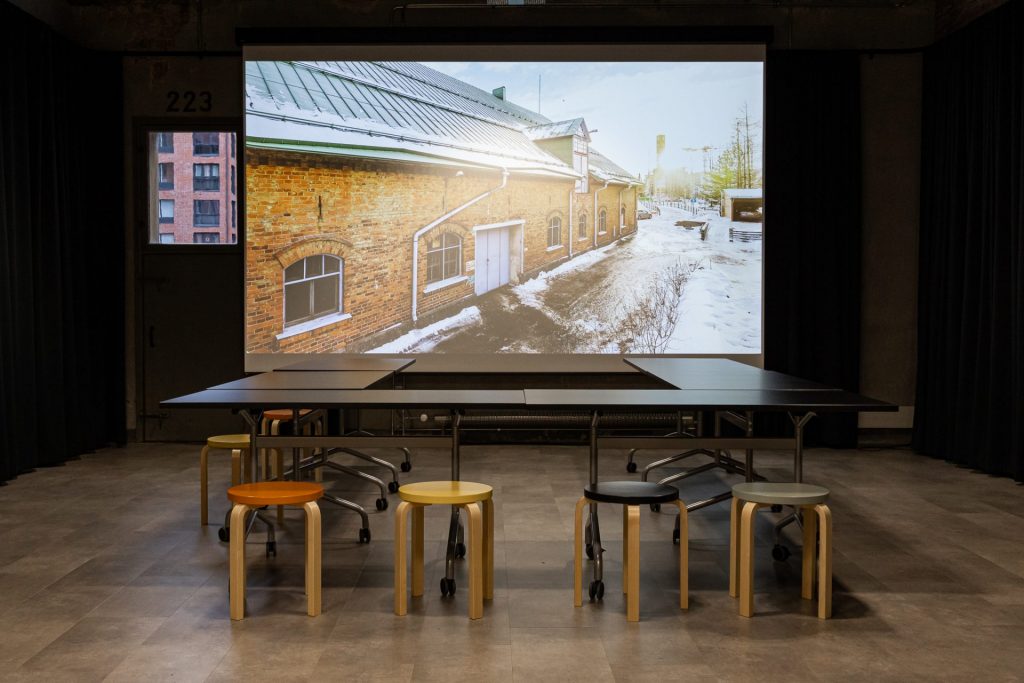 Workshop space that has chairs and tables, and in the background there is a picture of Kalevan Navetta on a screen.