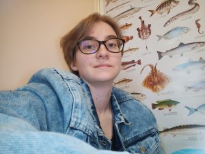 Nainen katsoo kameraan päin. puolikuva. Takana seinällä kalajuliste jossa monta eri kalaa.